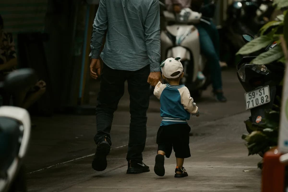 A father walks with a small child holding hands on a narrow city street with motorbikes.