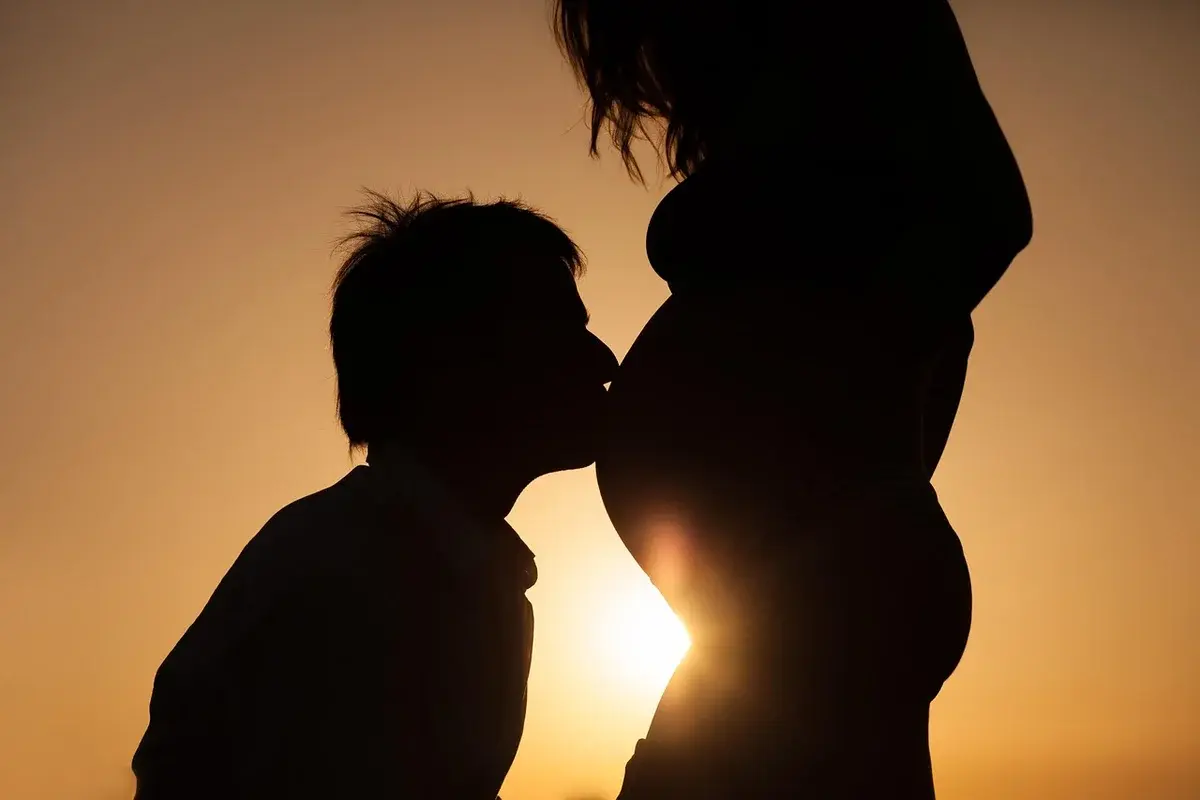 Silhouette of a man kissing a pregnant woman's belly at sunset.