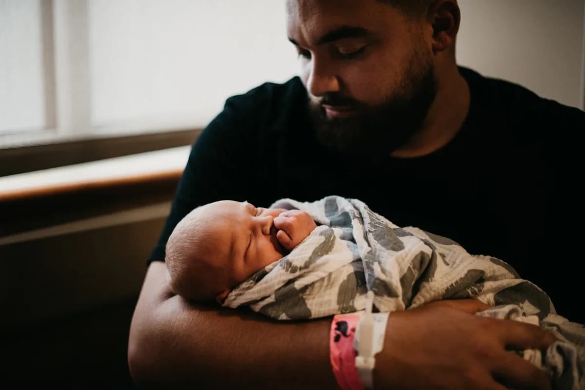 A bearded father holds a sleeping newborn wrapped in a patterned blanket near a window.
