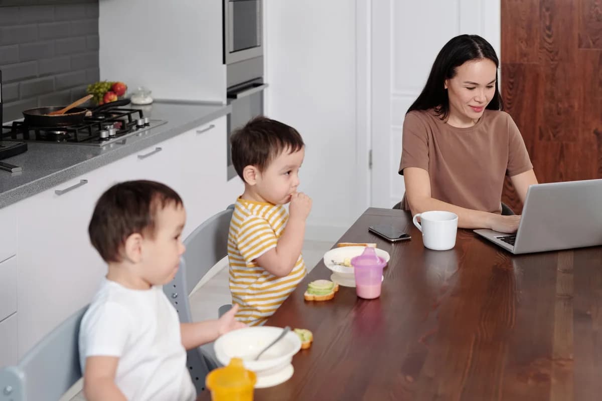 A mother uses a laptop at breakfast while two children eat at the table.