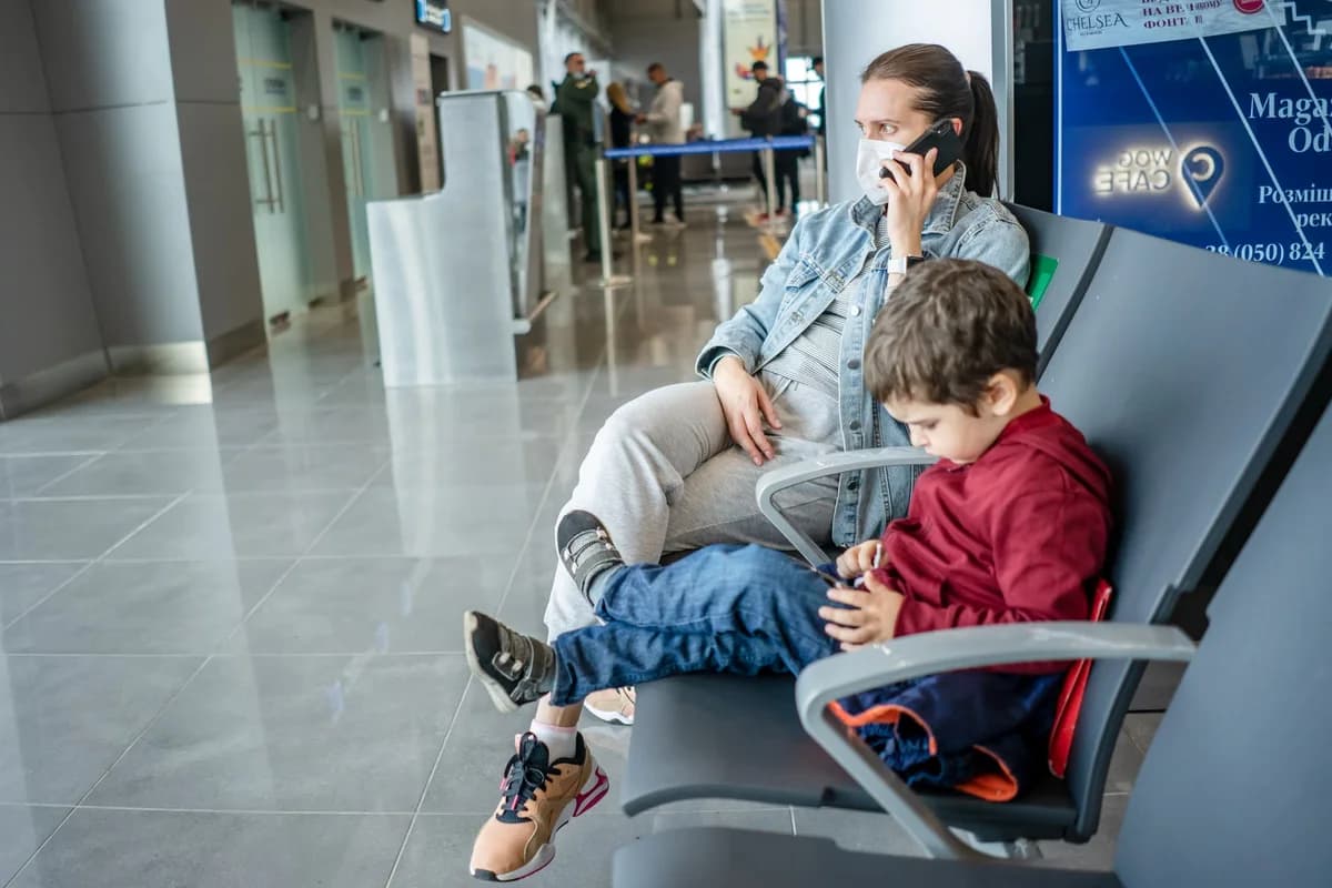 A masked mother talks on a phone while sitting next to a child using a device in an airport terminal.