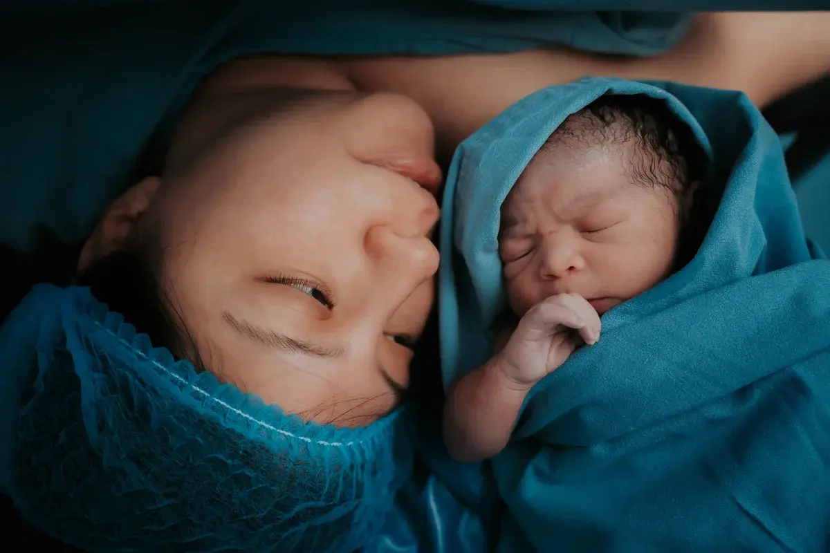 A mother lies next to her newborn baby, both wrapped in blue hospital blankets.
