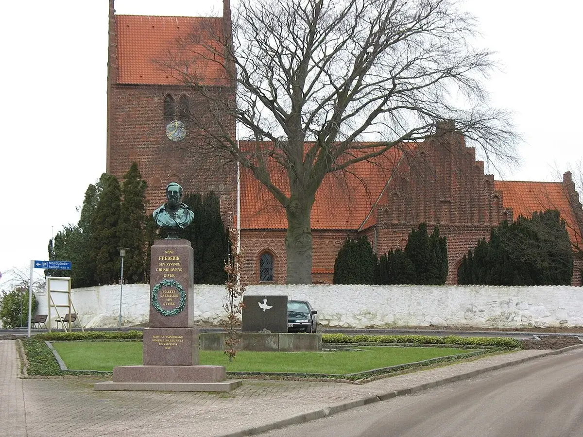 Brick church with a red roof behind a monument and trees.