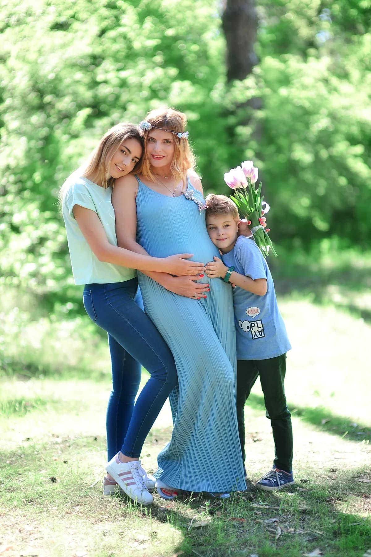 Pregnant mother posing with two children in a park, one holding flowers.