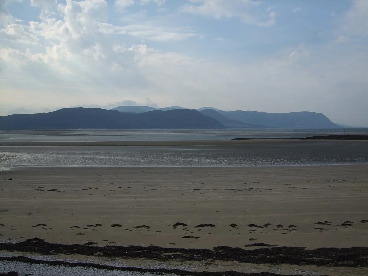 A sandy Welsh coastline with distant hills under a cloudy sky.